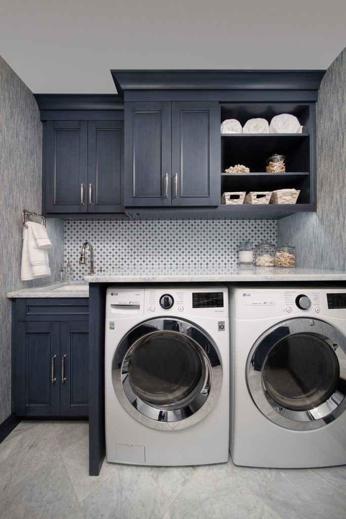 Compact laundry room with blue grey cabinets - small lower cabinet on left with sink, washer and dryer to right. White and blue tile backsplash, blue grey upper cabinets.