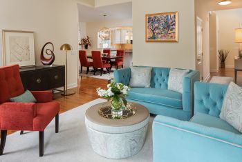 living room with light blue loveseat couches on right, white ottoman, and red-orange medium sized armchair on left, view into dining room