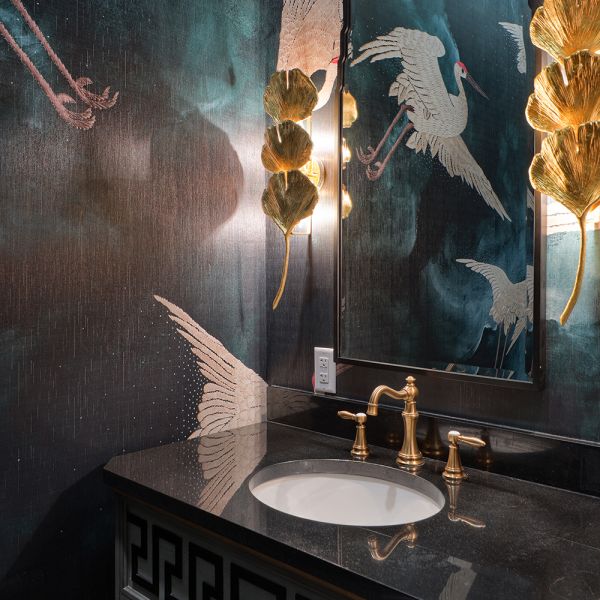 elegant powder room, flying cranes on moody teal and dark blue wallpaper, reflected in mirror with 3-tiered gold lotus leaf stack sconces on sides; furniture style vanity with black counter and gold faucet