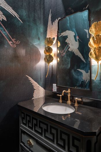 elegant powder room, flying cranes on moody teal and dark blue wallpaper, reflected in mirror with 3-tiered gold lotus leaf stack sconces on sides; furniture style vanity with black counter and gold faucet